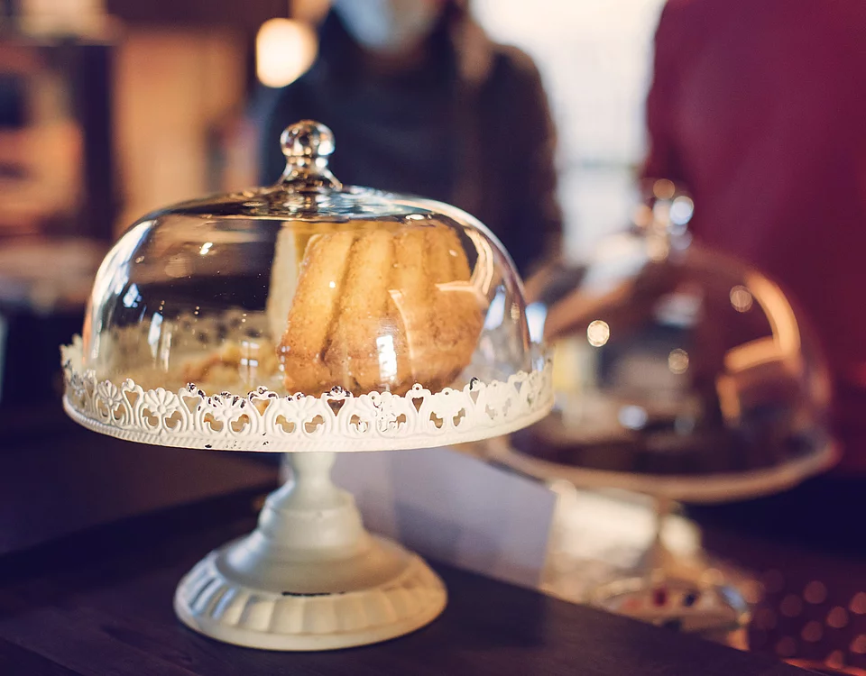 Eingedeckter Kuchenteller mit mehreren Keksen oder Gebäckstücken darunter, unscharf im Hintergrund Personen in einem Café.