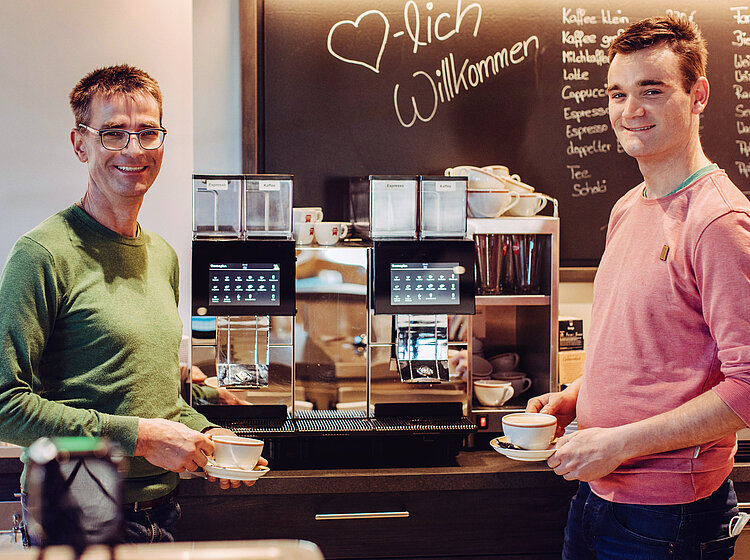 Zwei Männer stehen an einer Kaffeemaschine in einem Café, halten Kaffeetassen und lächeln in die Kamera; im Hintergrund eine Tafel mit der Aufschrift "Herzlich Willkommen".