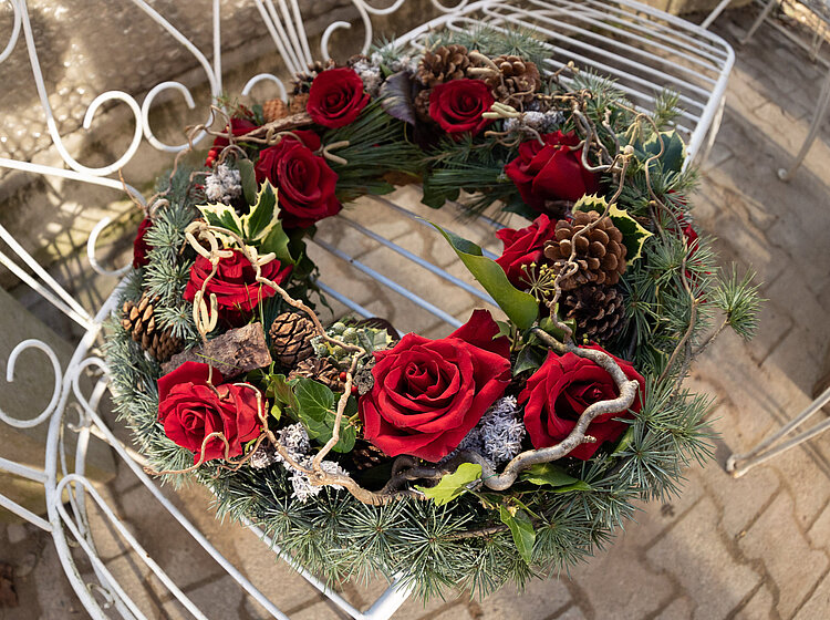 Weihnachtlicher Adventskranz aus Tannenzweigen, roten Rosen, Kiefernzapfen und dekorativen Ästen auf einem weißen Metallstuhl.
