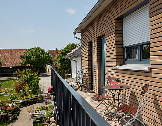 Balkon mit Holzwand, zwei Stühlen und Tisch, Blick auf Garten mit Pflanzen und Gebäuden im Hintergrund an sonnigem Tag.