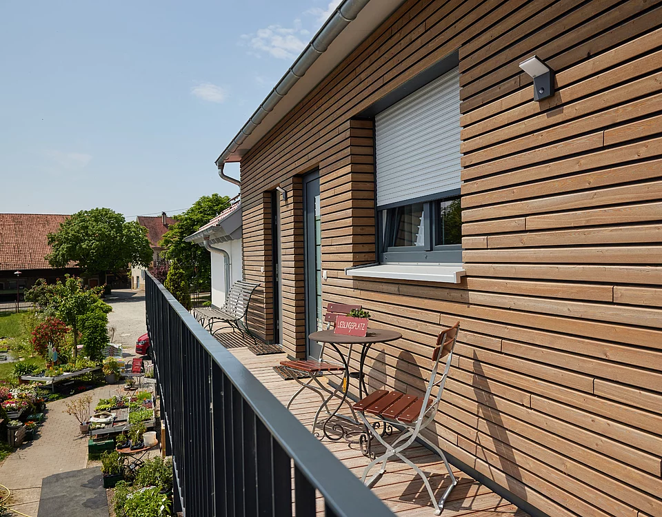 Balkon mit Holzwand, zwei Stühlen und Tisch, Blick auf Garten mit Pflanzen und Gebäuden im Hintergrund an sonnigem Tag.