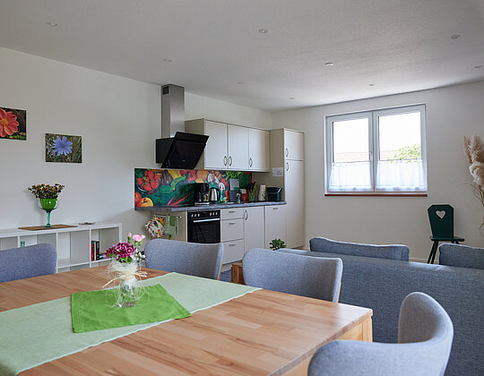 Offene Wohnküche mit Esstisch, grauen Stühlen, einer Küchenzeile mit buntem Fliesenspiegel und einem Fenster, dazu Blumendekoration auf dem Tisch und an der Wand.