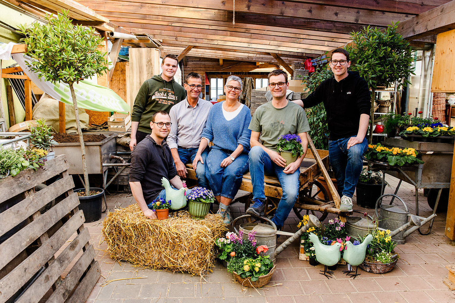 Sechs Personen in einer Gewächshaus-Umgebung umgeben von Blumen, Pflanzen und Gartengeräten, die gemeinsam lächelnd in die Kamera schauen.
