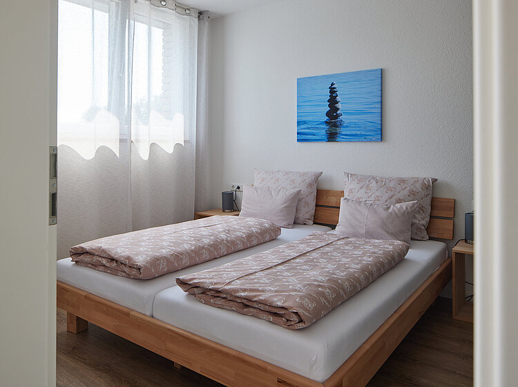 Schlicht eingerichtetes Schlafzimmer mit einem Holzrahmenbett, zwei beige Bettdecken und Kissen, einem Fenster mit weißen Gardinen und einem blauen Bild an der Wand.