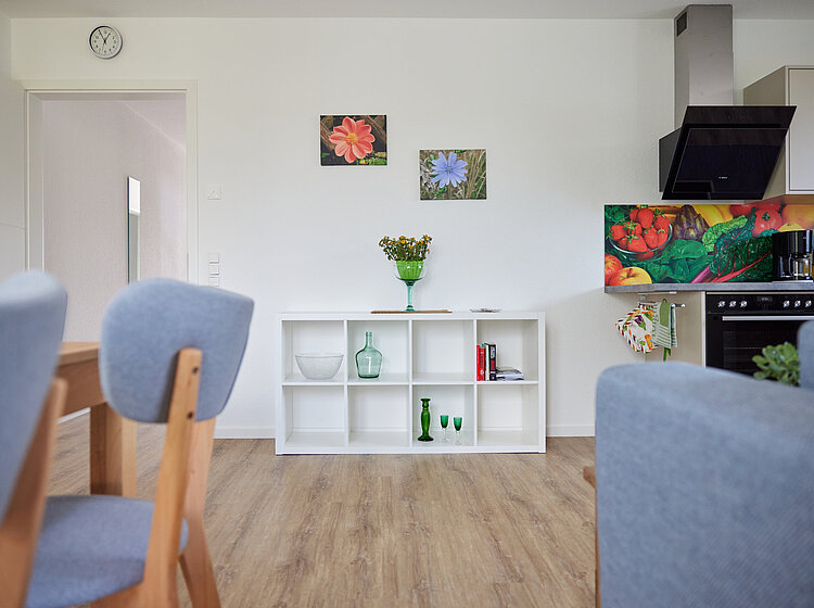 Moderner Wohn- und Essbereich mit Holzboden, grauen Stühlen, weißem Sideboard mit Deko und Blumen sowie bunter Küchenrückwand mit Gemüse-Motiv.