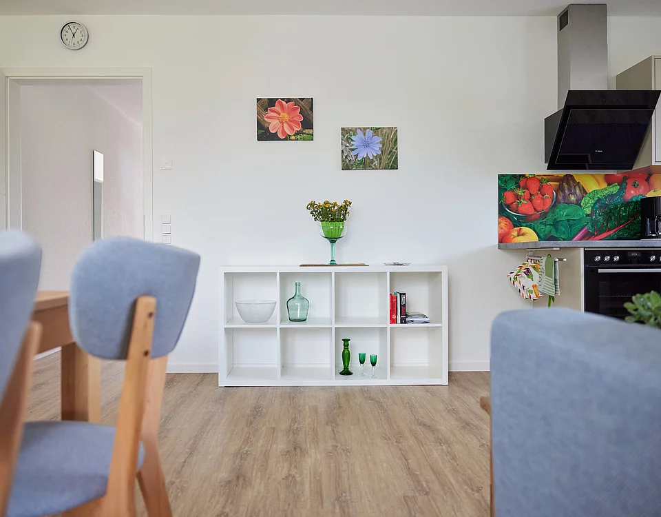 Moderner Wohn- und Essbereich mit Holzboden, grauen Stühlen, weißem Sideboard mit Deko und Blumen sowie bunter Küchenrückwand mit Gemüse-Motiv.