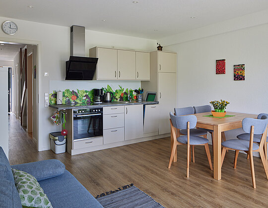 Moderner Wohnbereich mit offener Küche, Esstisch mit vier Stühlen, Sofa und Dekorationen an der Wand und auf der Arbeitsplatte.