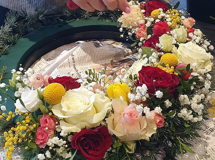 Person bindet einen bunten Blumenkranz mit roten, weißen und rosa Rosen sowie anderen kleinen Blumen und Grünzeug.
