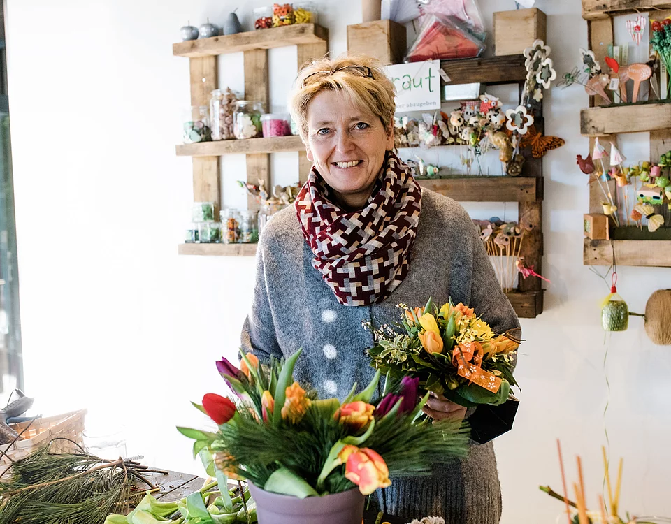 Frau in einem Blumenladen hält einen Blumenstrauß und steht vor einem Tisch mit Pflanzen und Blumenarrangements. Im Hintergrund sind dekorative Regale mit weiteren Blumen und Dekorationsartikeln zu sehen.