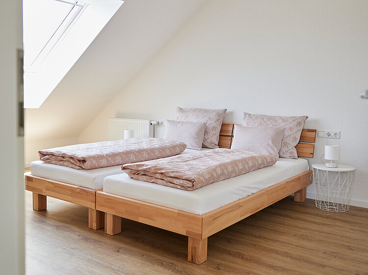 Schlicht eingerichtetes Schlafzimmer mit zwei Einzelbetten aus Holz, rosafarbenen Bettdecken und Kissen, einem Dachfenster und hellem Holzboden.
