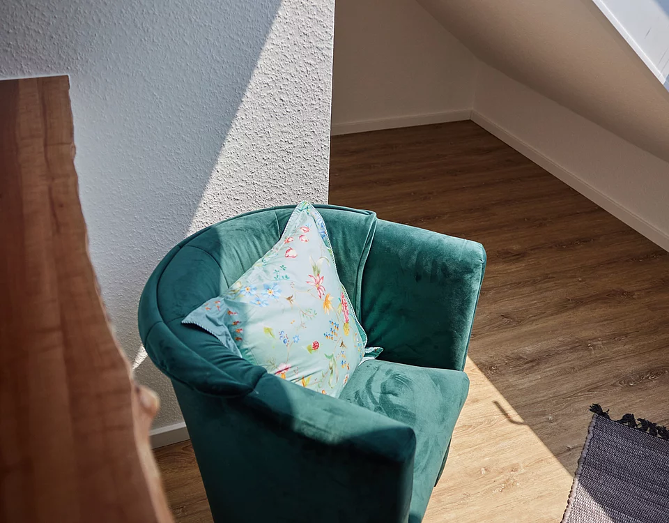 Grüner Sessel mit floralem Kissen steht in hellem Raum mit Dachschräge und Holzfußboden, Sonnenlicht fällt auf den Stuhl.