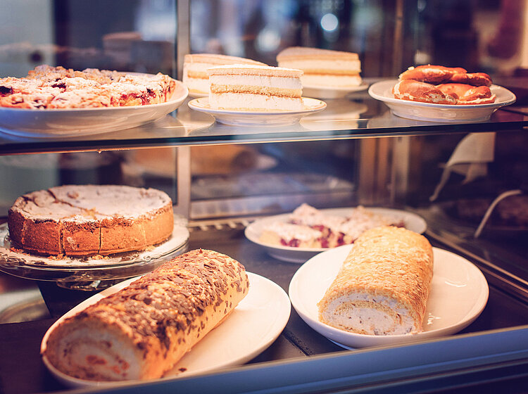 Vitrine mit verschiedenen Kuchen und Gebäckstücken wie Blechkuchen, Sahneschnitten und Rouladen auf Tellern.
