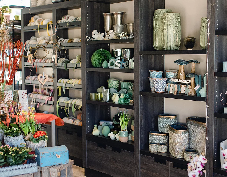 Regal mit verschiedenen Wohnaccessoires und Keramikartikeln sowie links Blumen und Dekorationsgegenstände in einem Geschäft.