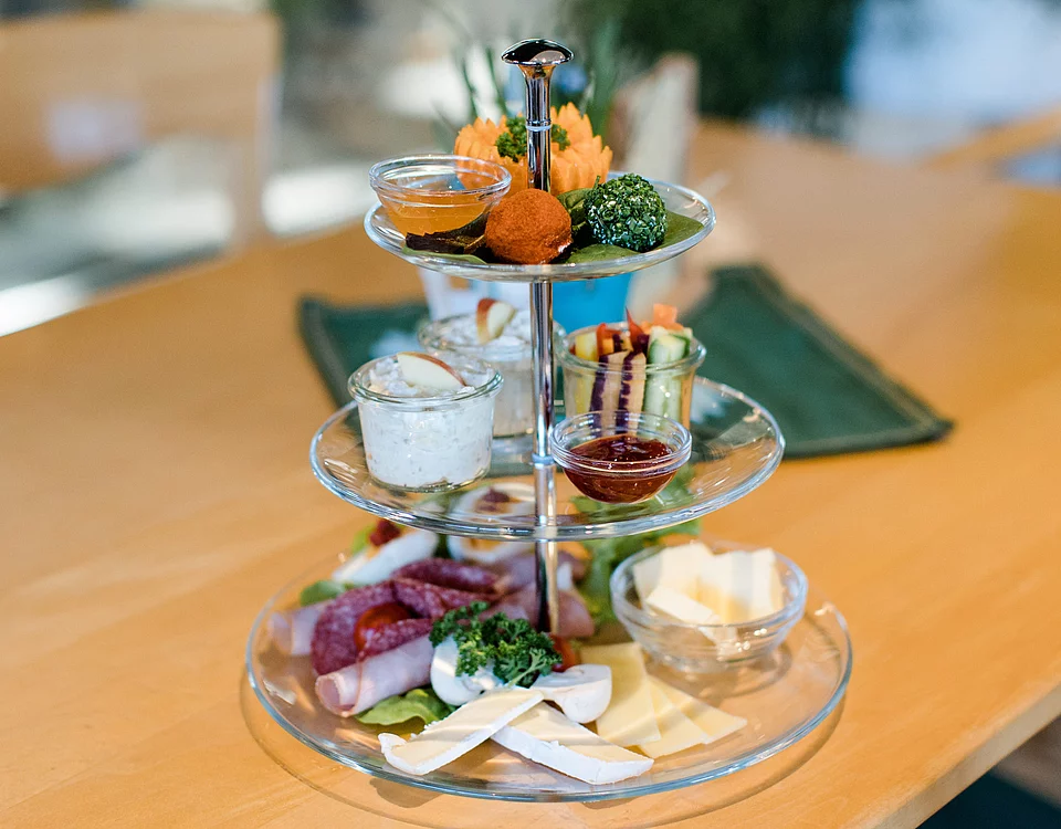 Dreistöckiges Glas-Etagere mit verschiedenen Käsesorten, Wurst, Frischkäse, Dips und dekorativen herzhaften Snacks auf einem Holztisch.