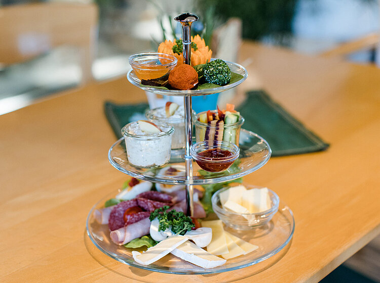 Dreistöckiges Glas-Etagere mit verschiedenen Käsesorten, Wurst, Frischkäse, Dips und dekorativen herzhaften Snacks auf einem Holztisch.