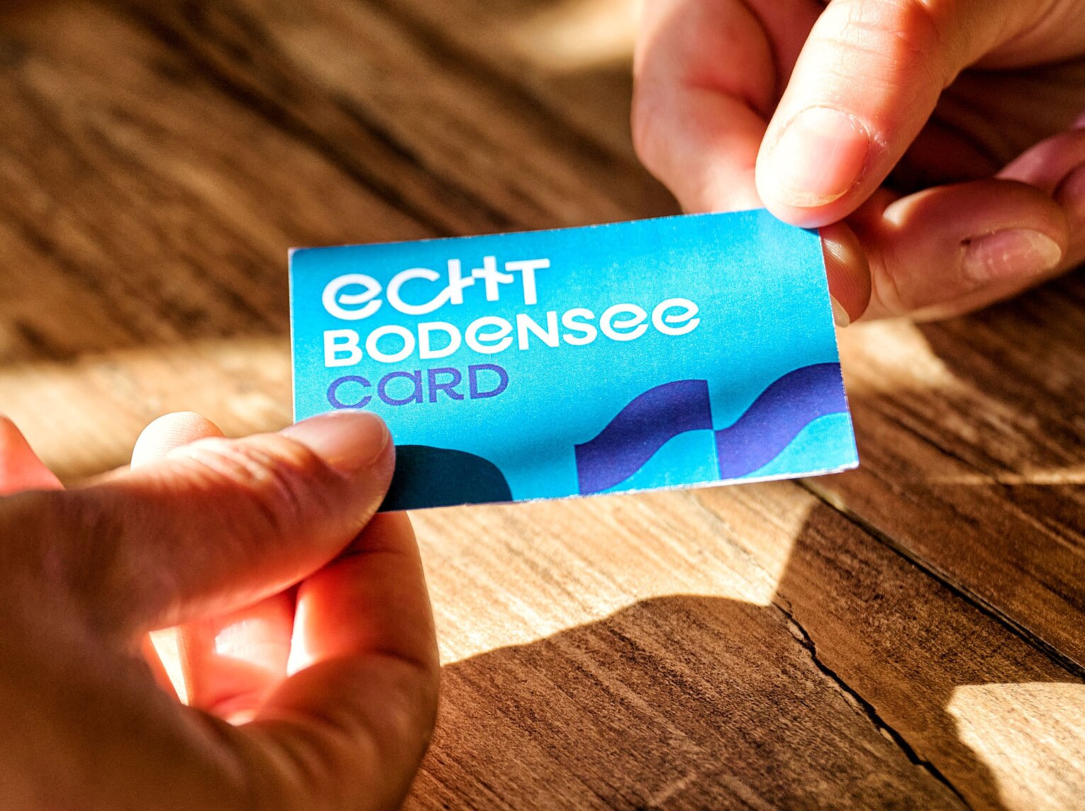 Zwei Hände halten eine blaue Karte mit der Aufschrift echt Bodensee Card über einem Holztisch.