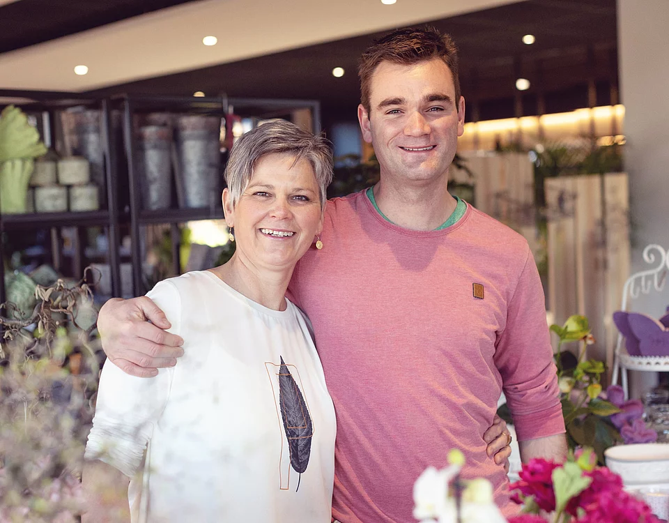 Zwei lächelnde Personen umarmen sich in einem hellen Raum mit Blumen und Pflanzen im Hintergrund.