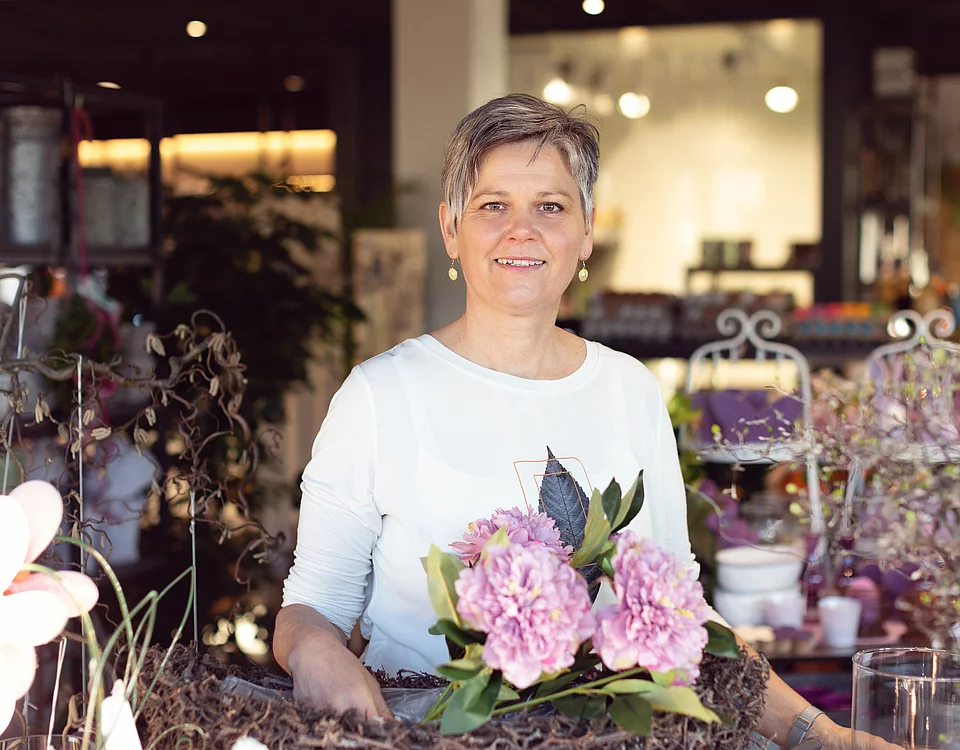 Frau in weißem Oberteil steht in einem Blumenladen und hält einen Blumenstrauß mit rosa Hortensien.