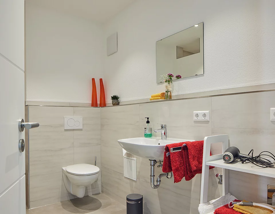 Modernes Badezimmer mit weißer Wand, Wandtoilette, Waschbecken, Spiegel und einem kleinen Regal mit Handtüchern und Haartrockner.