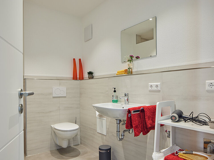 Modernes Badezimmer mit weißer Wand, Wandtoilette, Waschbecken, Spiegel und einem kleinen Regal mit Handtüchern und Haartrockner.