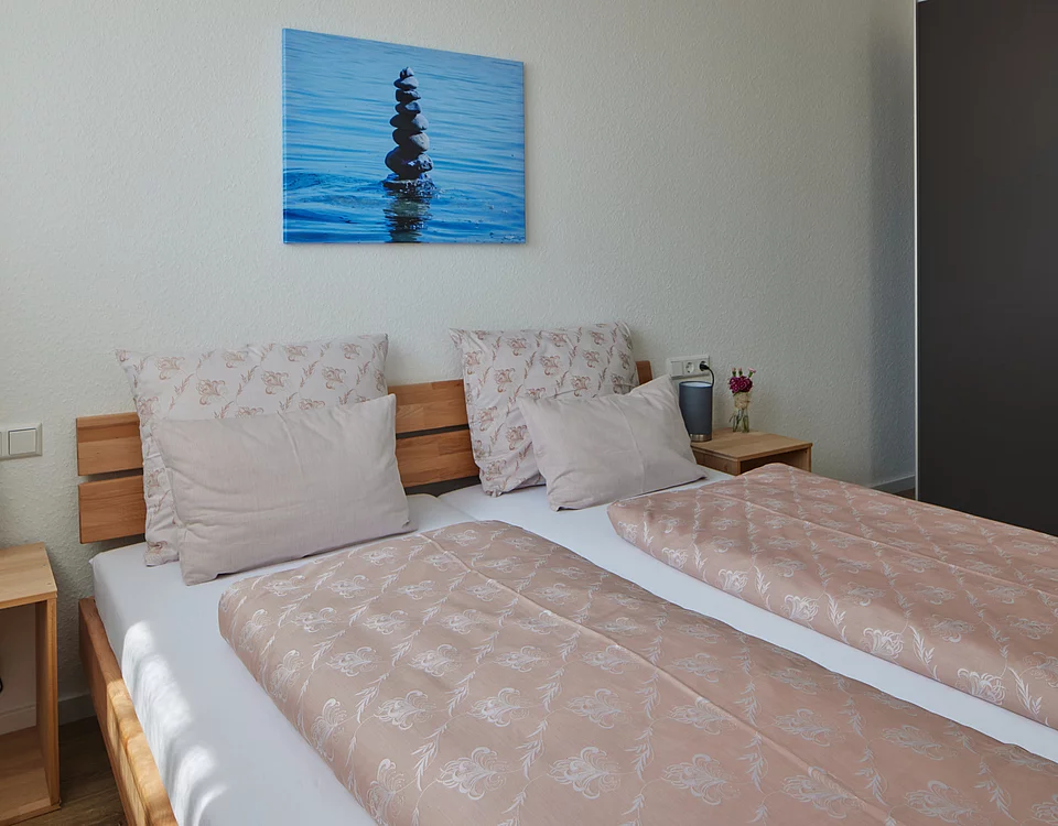 Schlicht eingerichtetes Schlafzimmer mit zwei Einzelbetten, beigen Bettdecken und Kissen, zwei Nachttischen mit Lampe und Blume sowie einem Bild von einem Steinturm auf Wasser an der Wand.