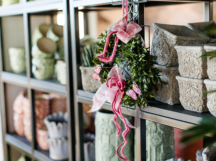 Regal mit verschiedenen dekorativen Blumenkübeln, darunter ein grüner Kranz mit pinken Bändern und Schmetterlingen als Deko hängen im Vordergrund.