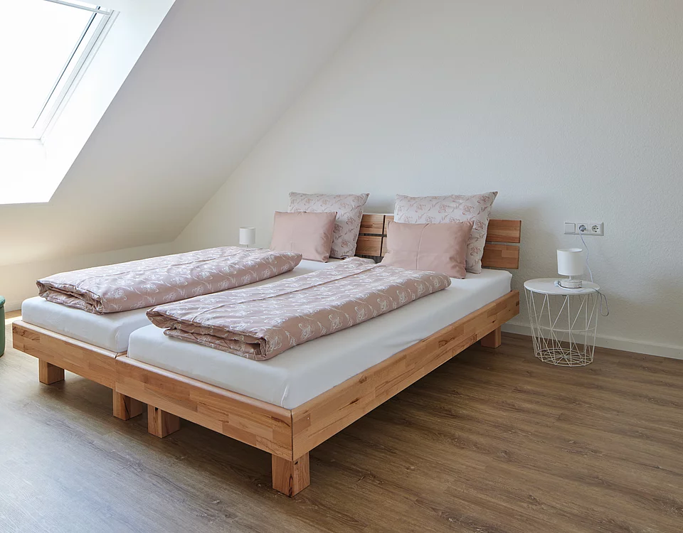 Helles Schlafzimmer mit zwei Einzelbetten aus Holz, rosa Bettwäsche und Kissen, kleinem weißen Nachttisch und Holzboden.