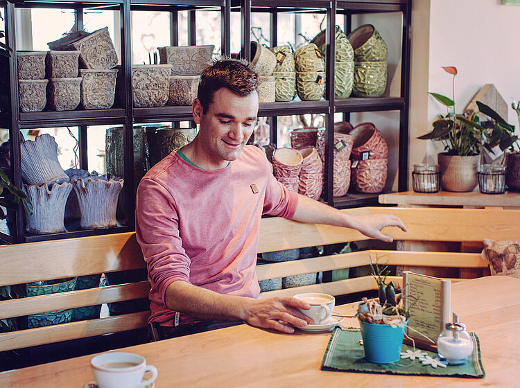 Ein junger Mann sitzt an einem Holztisch in einem gemütlichen Café und hält eine Tasse Kaffee, umgeben von Dekorationen und Pflanzen.
