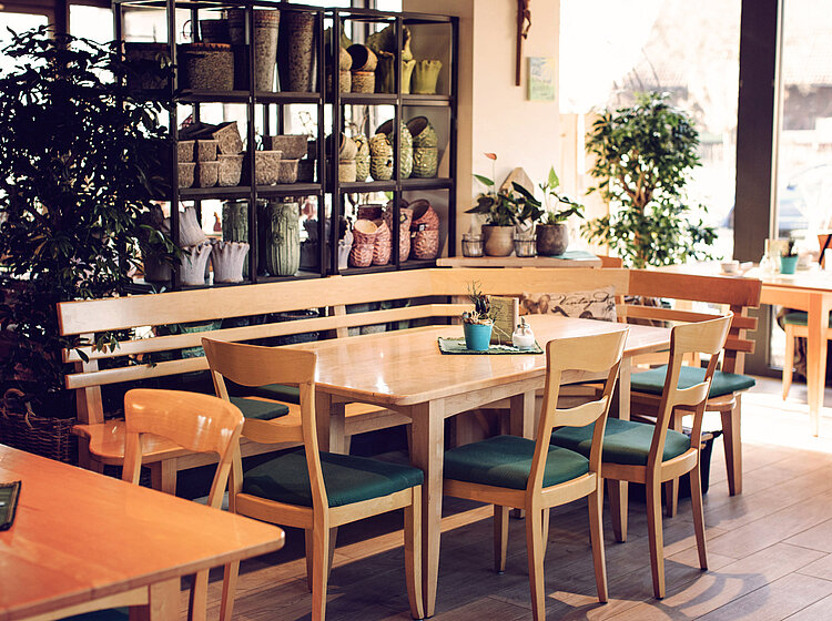 Innenansicht eines Cafés mit Holztischen, Stühlen mit grünen Sitzpolstern und Regalen mit Körben und Pflanzen im Hintergrund.