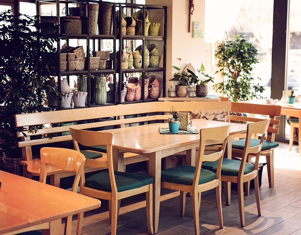 Innenansicht eines Cafés mit Holztischen, Stühlen mit grünen Sitzpolstern und Regalen mit Körben und Pflanzen im Hintergrund.