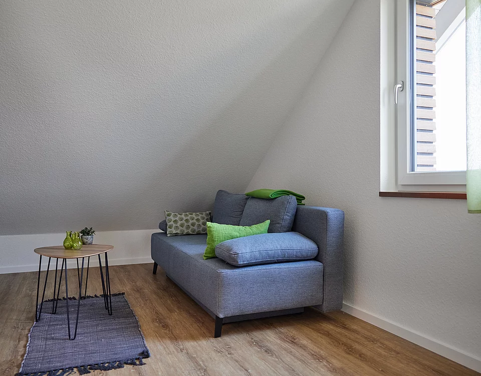 Kleines Wohnzimmer mit grauem Sofa, bunten Kissen, runden Holztisch mit Pflanzen und offenem Fenster mit grüner Gardine.