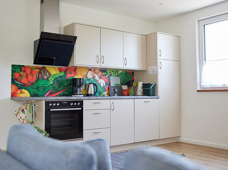 Moderne Küchenzeile mit weißen Schränken, buntem Gemüse-Motiv als Fliesenspiegel, Herd, Kaffeemaschine und Wasserkocher, daneben ein Fenster mit weißem Vorhang.