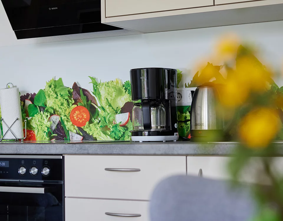 Moderne Küchenzeile mit Kaffeemaschine, Wasserkocher und einem bunten Gemüse-Motiv als Spritzschutz an der Wand.