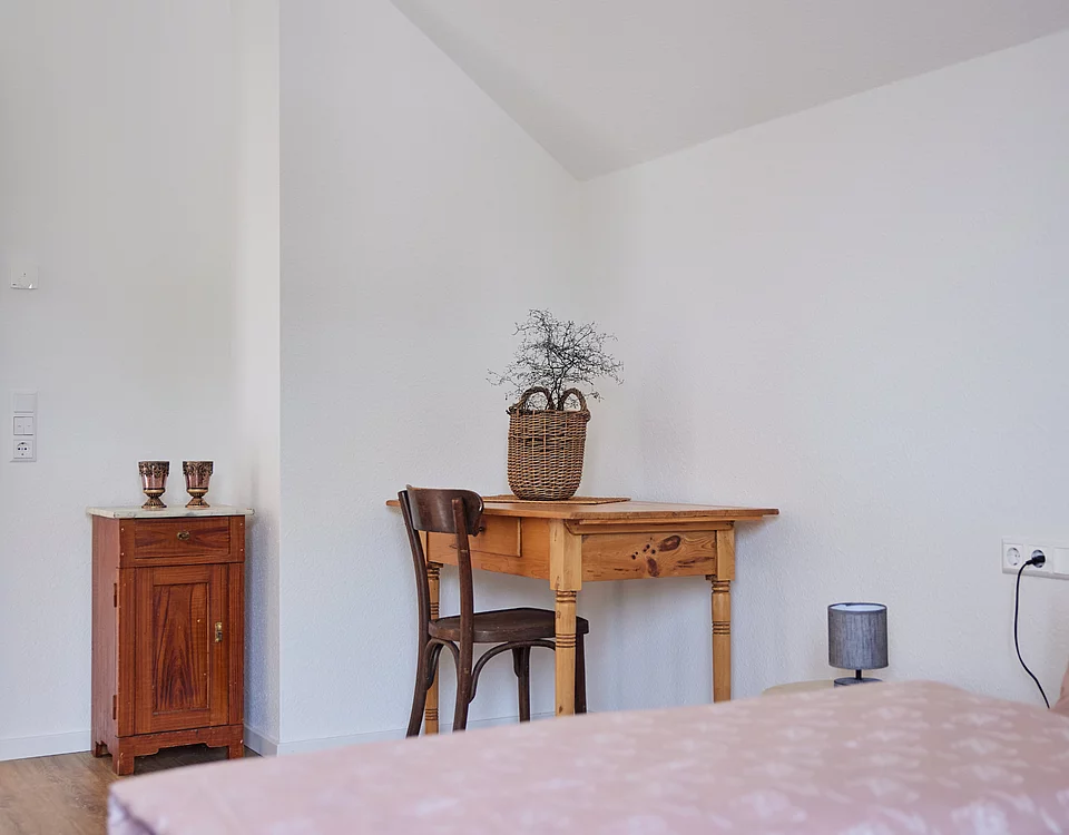 Schlicht eingerichtetes Zimmer mit Holztisch, Stuhl, Korb mit Zweigen, kleinem Nachttisch und einem Bett mit rosa Bettdecke.