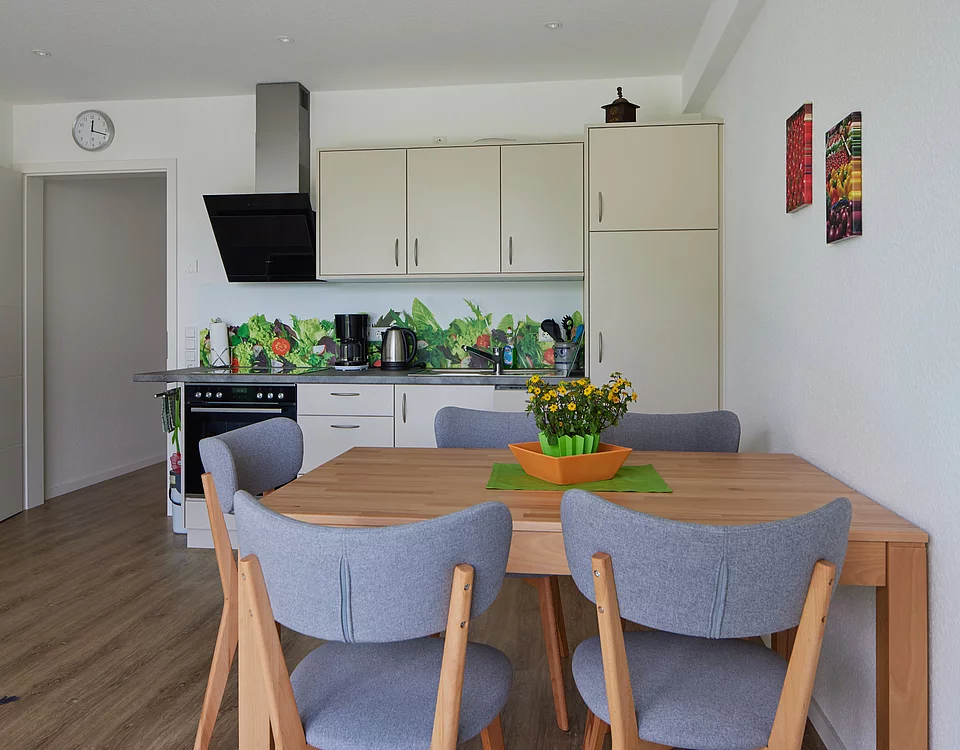 Moderner Küchen- und Essbereich mit Holzfußboden, hellem Holztisch, grauen Stühlen, weißen Küchenschränken und buntem Gemüse-Motiv auf der Küchenrückwand.