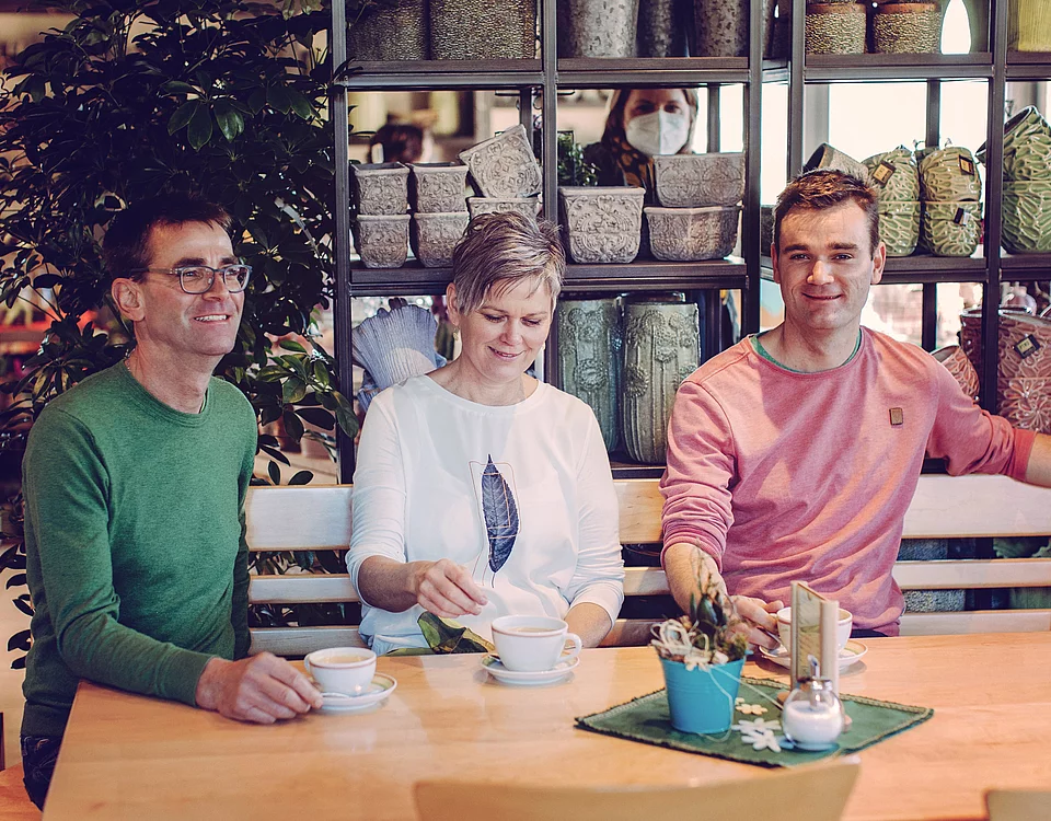 Drei Personen sitzen entspannt an einem Holztisch in einem Café, halten Tassen und lächeln. Im Hintergrund sind Regale mit Körben und Pflanzen zu sehen.