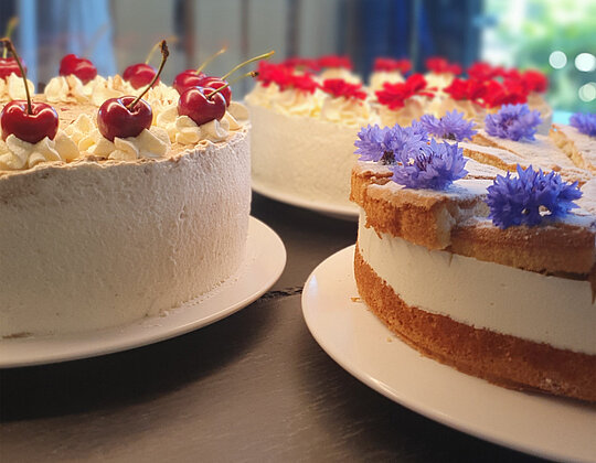Drei verschiedene dekorierte Kuchen in einer Vitrine, einer mit Kirschen, einer mit roten Blumen und einer mit blauen Blüten.
