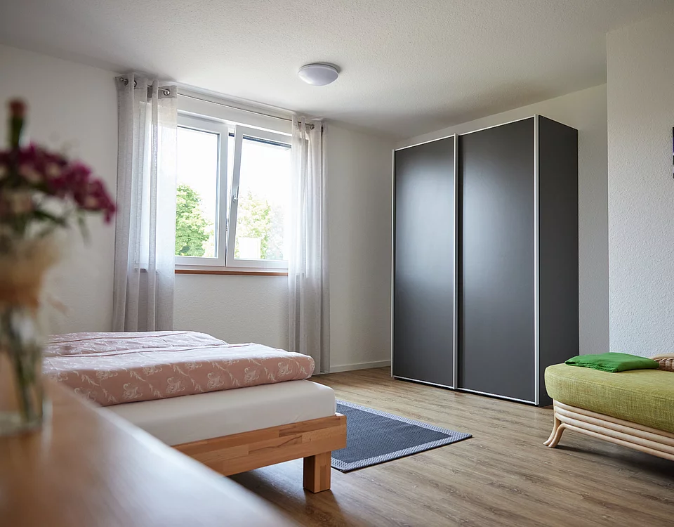 Helles Schlafzimmer mit einem Bett, einem großen Fenster mit weißen Vorhängen, einem schwarzen Kleiderschrank und einem grünen Sofa. Holzboden und frische Blumen auf einem Tisch im Vordergrund.