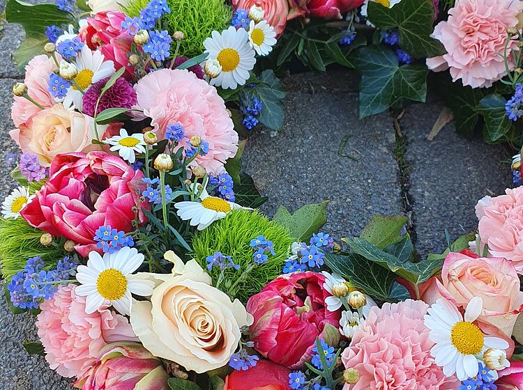 Blumenkranz aus verschiedenen bunten Blumen wie Rosen, Nelken, Gänseblümchen und grünen Blüten auf grauem Steinboden.