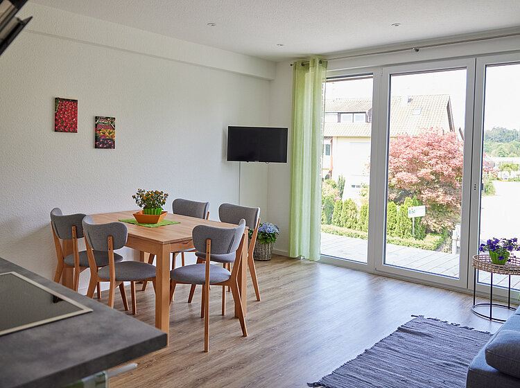 Heller Essbereich mit Holztisch und vier Stühlen neben großen bodentiefen Fenstern mit grünlichen Vorhängen, Blick auf Garten mit Baum und umliegende Häuser.