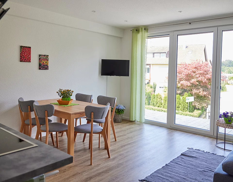 Heller Essbereich mit Holztisch und vier Stühlen neben großen bodentiefen Fenstern mit grünlichen Vorhängen, Blick auf Garten mit Baum und umliegende Häuser.