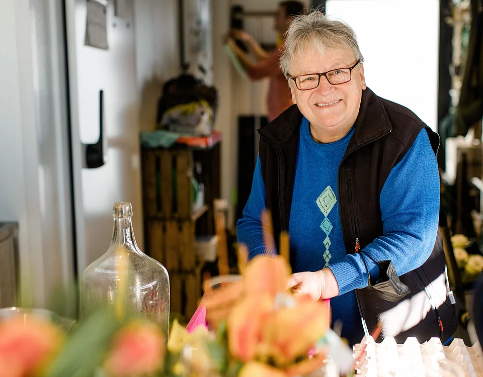 Älterer Mann mit Brille und blauer Kleidung lächelt in einem hellen Raum mit Pflanzen und Dekorationsartikeln.