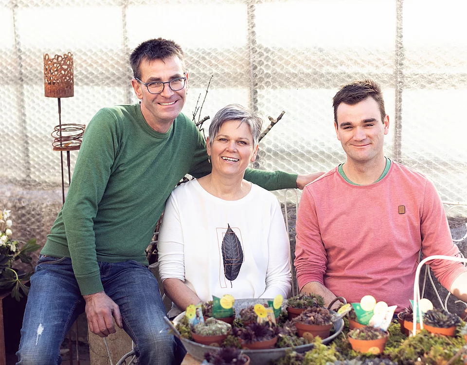 Drei Personen sitzen zusammen in einem Garten oder Gewächshaus, umgeben von Töpfen mit Pflanzen und Blumen, und lächeln in die Kamera.