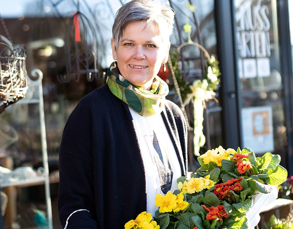 Frau mit kurzem Haar steht draußen vor einem Geschäft und hält einen Blumentopf mit gelben und roten Primeln.