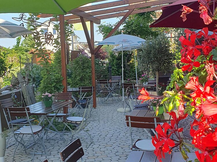 Leerer Außenbereich eines Cafés mit Tischen, Stühlen, bunten Sonnenschirmen und roten Blumen im Vordergrund bei sonnigem Wetter.