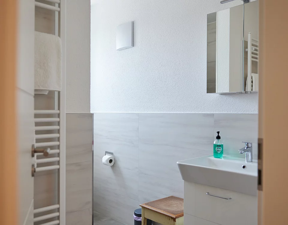 Blick in ein modernes Badezimmer mit Waschbecken, Spiegelschrank, Handtuchhalter und Holzhocker.