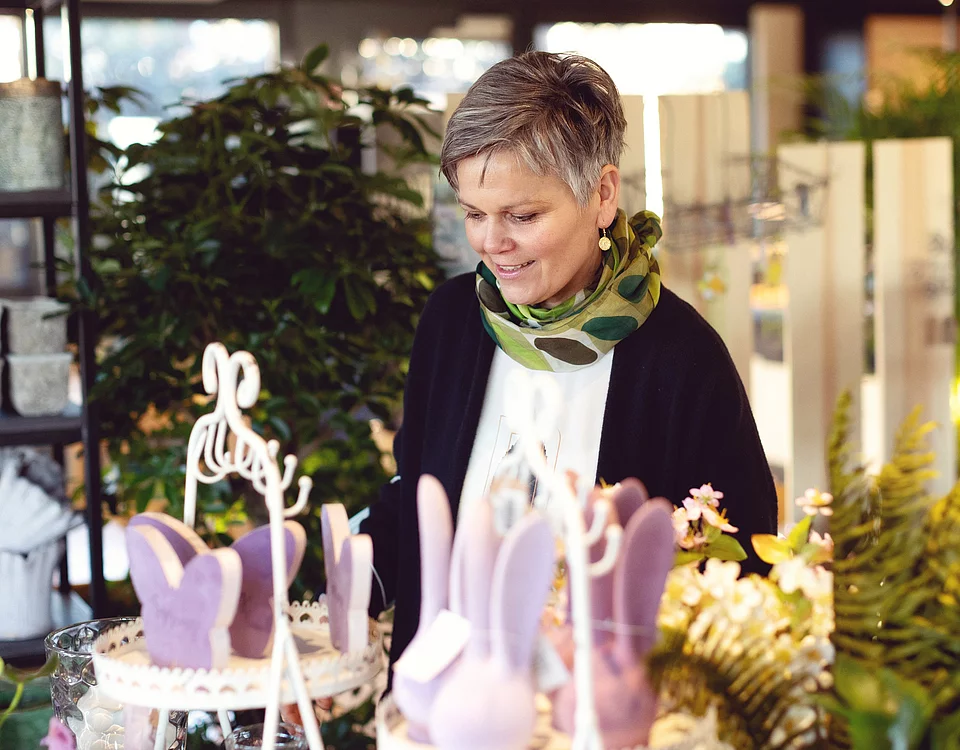Frau mit kurzem Haar schaut lächelnd auf bunte Osterdekoration in einem Laden mit Pflanzen und frühlingshaften Motiven.