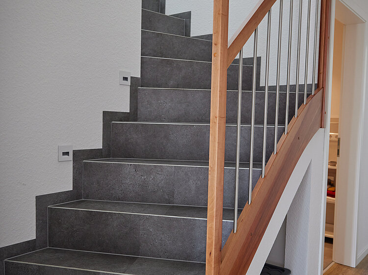Innertreppe mit grauen Fliesenstufen und Holz-Geländer in einem modernen Wohnhaus, mit weiß gestrichenen Wänden und Holzboden.