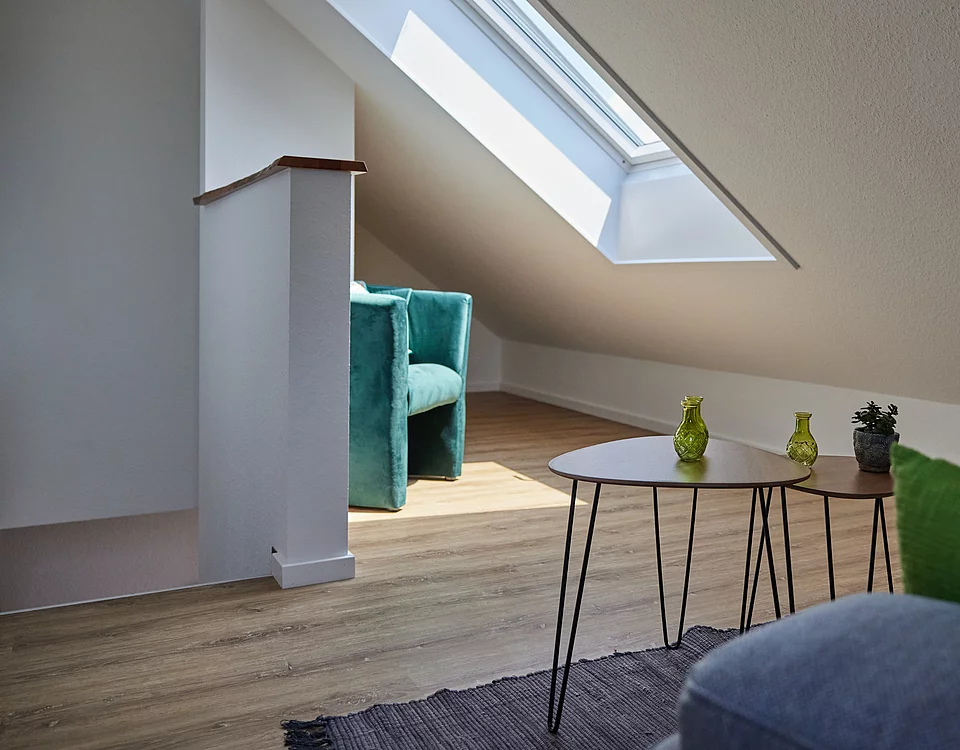 Helles Dachzimmer mit Dachfenster, einem grünen Sessel, zwei kleinen Tischen mit Dekoration und einem Teil eines Sofas.