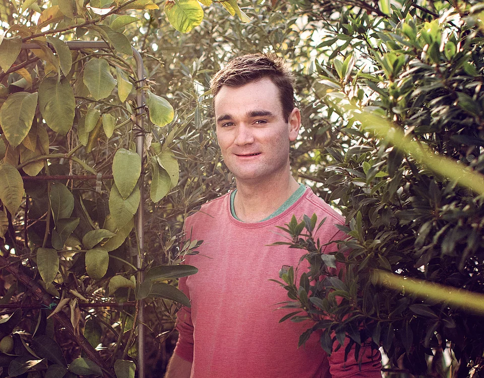 Mann in rotem Pullover steht lächelnd zwischen dichtem Grün von Bäumen und Sträuchern.